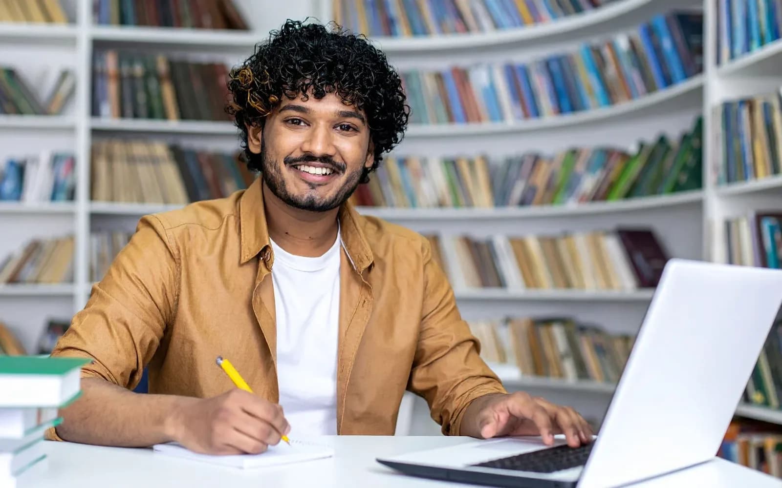 An adult taking notes beside a laptop in a library
